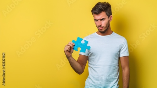 Male in t-shirt and jeans with a thoughtful expression holding a blue puzzle piece against a solid yellow background, capturing concentration in a minimalist setting