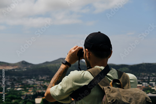Travel and adventure concept. Young man with a retro-style green backpack stays in old town Bar in Montenegro country. Guy Looking at nature and houses with tiled roofs in mountains using binoculars.