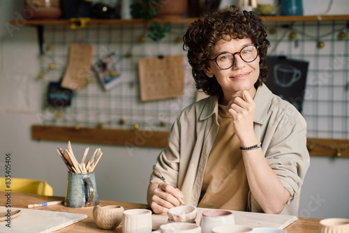 Confident female artisan wearing eyeglasses sitting at table in workshop with carving tool in hand looking at camera photo