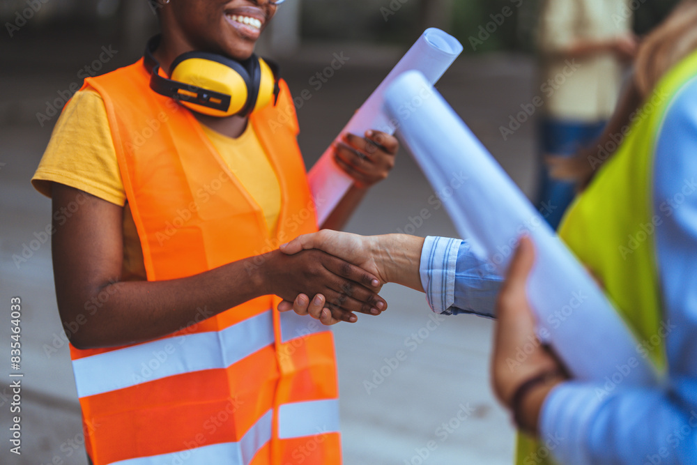 Two female construction professionals in reflective vests and safety gear shake hands at a building site, exchanging blueprints and collaboration. Two female construction professionals in reflective vests and safety gear shake hands at a building site, exchanging blueprints and collaboration.