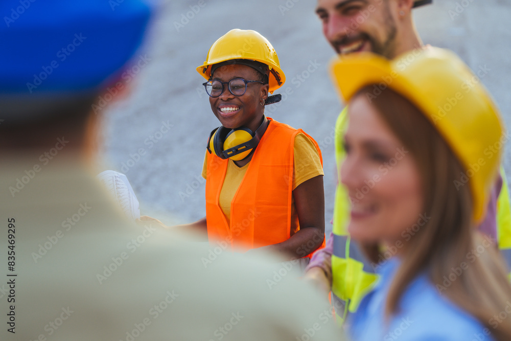 A group of smiling diverse construction professionals, wearing safety helmets and vests, engage in planning at a construction site. A group of smiling diverse construction professionals, wearing safety helmets and vests, engage in planning at a construction site.