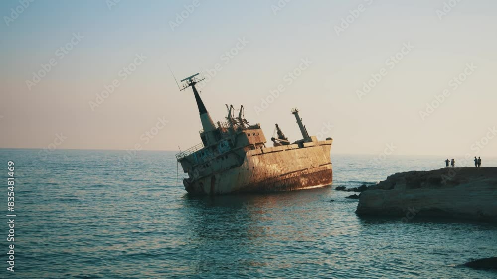 An abandoned shipwreck rests near coast, drawing attention of onlookers and tourists on a rocky outcrop, as calm sea under a blue sky.