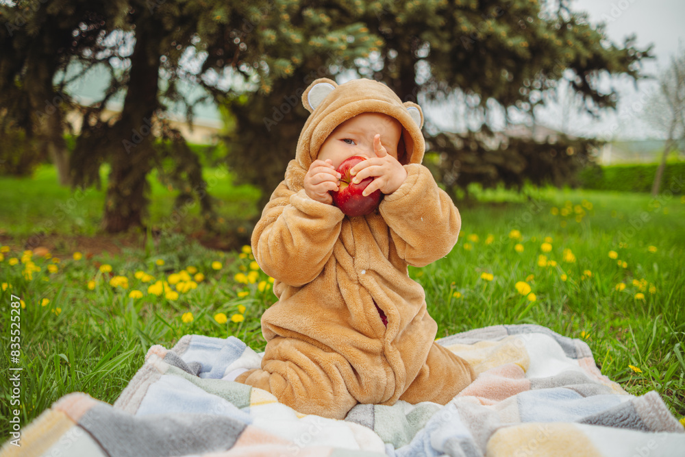 Cute baby girl in plush suit with red apple in hands sitting on blanket in green grass. Cute baby girl in plush suit with red apple in hands sitting on blanket in green grass.