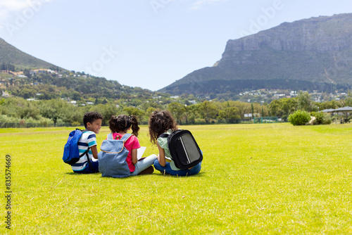 Three biracial children sit on a grassy field, backpacks on and facing away, with copy space