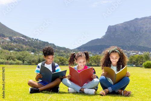 Biracial children are engrossed in reading books outdoors with copy space