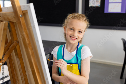 Caucasian girl with blonde hair is painting on an easel in school, wearing a colorful apron