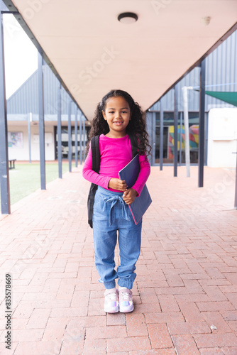 In school, outdoors, a biracial young girl holding a tablet stands smiling © WavebreakMediaMicro