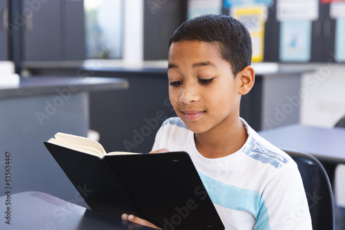In school, in the classroom, a young biracial student is engrossed in reading a book