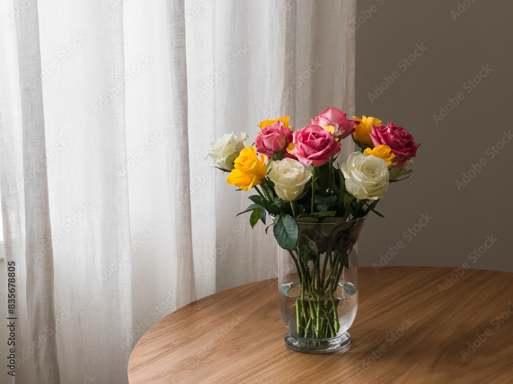 A bright bouquet of yellow, white, pink roses in a glass vase on a wooden table in the living room A bright bouquet of yellow, white, pink roses in a glass vase on a wooden table in the living room