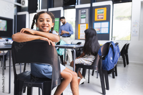 Biracial girl with braided hair smiles, sitting in a school classroom with copy space photo