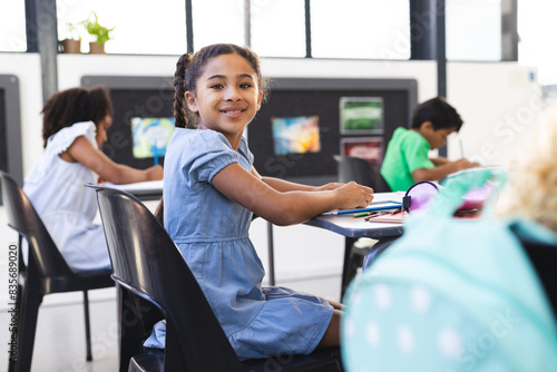 In school, young biracial girl with curly brown hair is sitting, smiling at camera photo