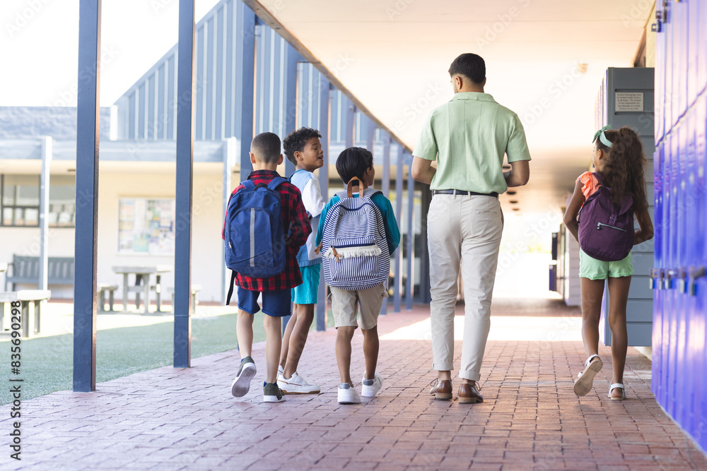 Young Asian man, a teacher, leads three biracial children through a school corridor Young Asian man, a teacher, leads three biracial children through a school corridor
