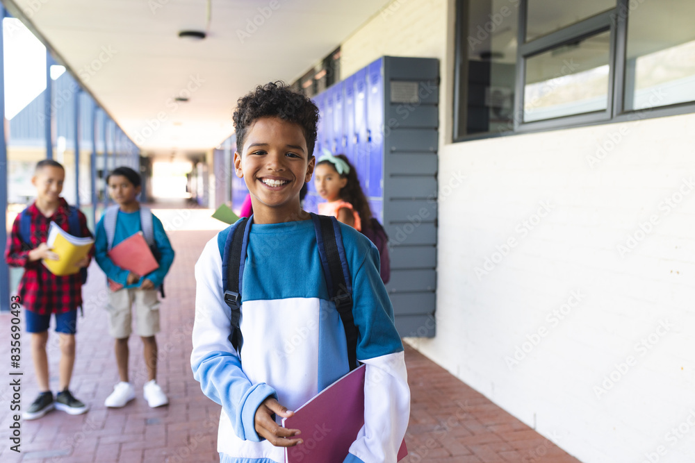 Biracial boy with a bright smile stands in a school hallway, holding a ...