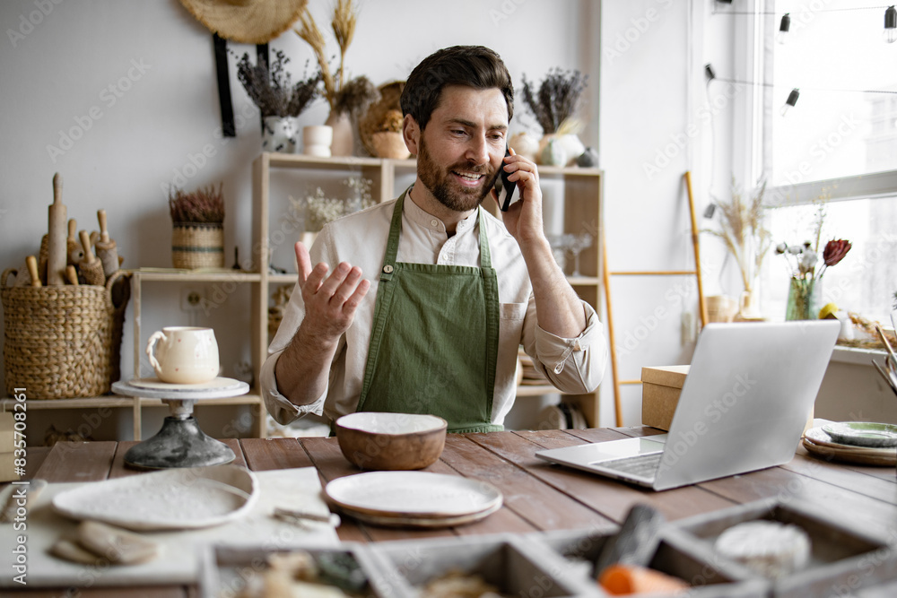 Caucasian male potter entrepreneur talking to buyer of handmade bowl sitting at table and taking order using laptop. Successful businessman owner of shop made of clay talking to customer. Caucasian male potter entrepreneur talking to buyer of handmade bowl sitting at table and taking order using laptop. Successful businessman owner of shop made of clay talking to customer.