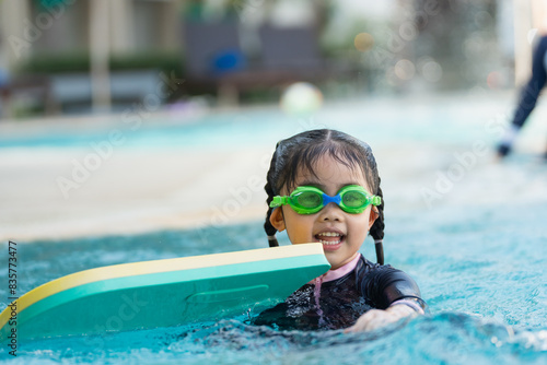 A young girl is in the water with a green and yellow boogie board. She is wearing a pink and black swimsuit and is smiling © Woraphon