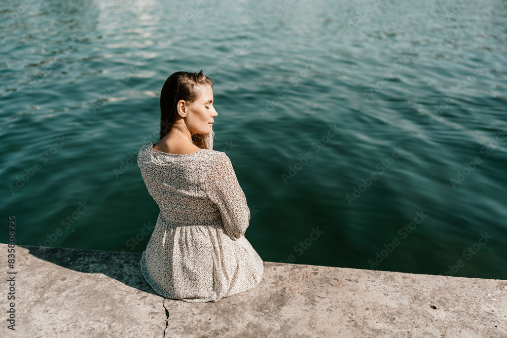 A woman is sitting on a ledge by a body of water. She is wearing a white dress and she is looking out at the water. The scene is peaceful and serene, with the woman taking a moment to enjoy the view. A woman is sitting on a ledge by a body of water. She is wearing a white dress and she is looking out at the water. The scene is peaceful and serene, with the woman taking a moment to enjoy the view.