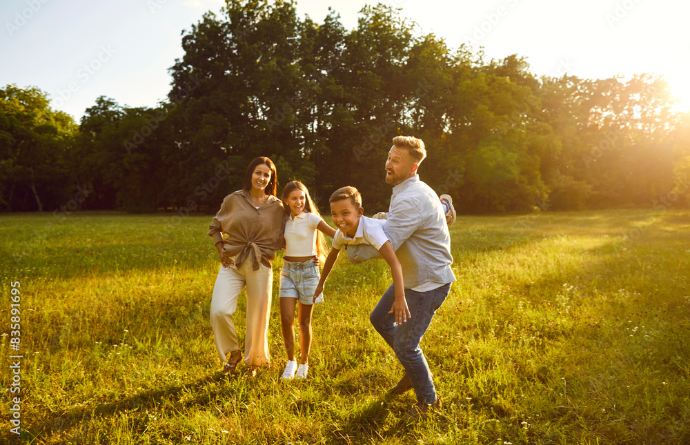 Happy young family of four having fun and playing plane at sunny day in nature. Parents mother, father and their children boy and girl walking in the field outdoors at sunset together. Happy young family of four having fun and playing plane at sunny day in nature. Parents mother, father and their children boy and girl walking in the field outdoors at sunset together.