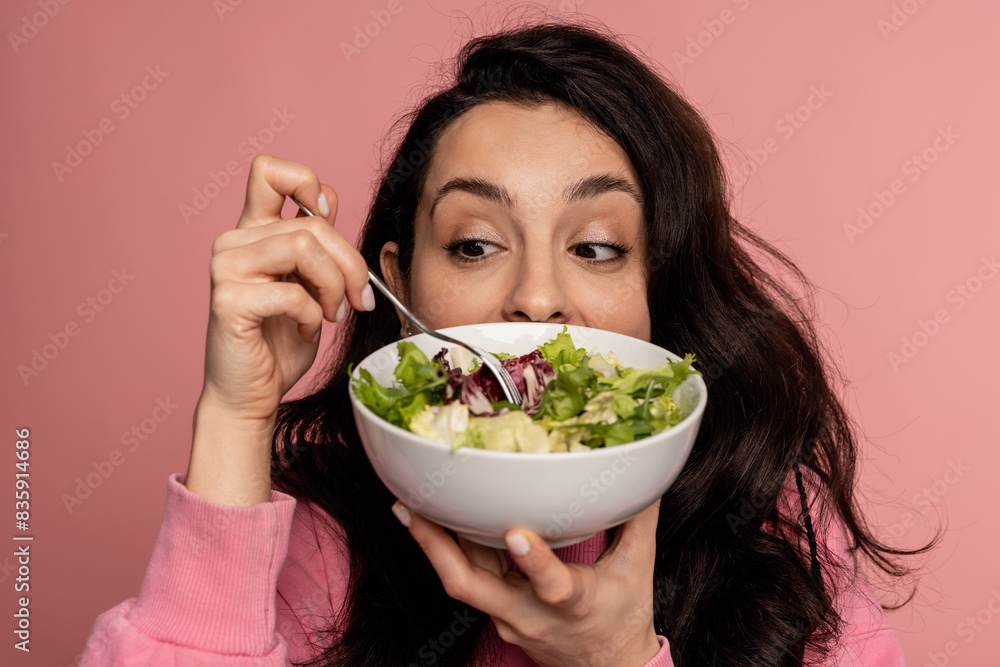 Young dark-haired woman eating a vegetarian dish in front of the camera on the pink background. Healthy diet concept Young dark-haired woman eating a vegetarian dish in front of the camera on the pink background. Healthy diet concept