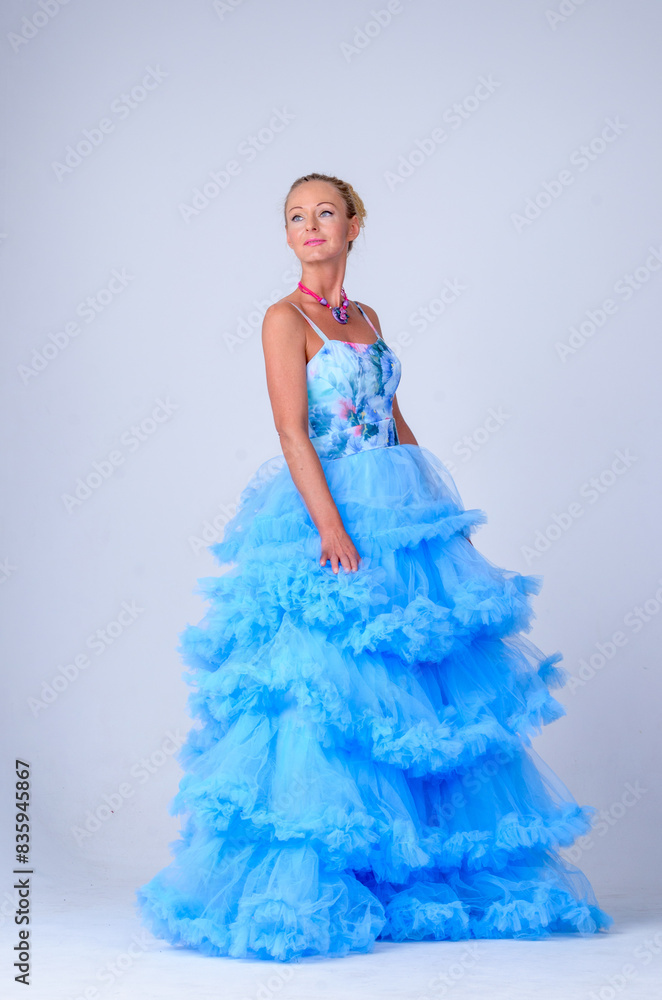 A beautiful woman in a blue dress with a red necklace poses in front of the camera. The dress is long and layered, giving it a flowy and elegant look. The woman's blond hair is styled in a bun. A beautiful woman in a blue dress with a red necklace poses in front of the camera. The dress is long and layered, giving it a flowy and elegant look. The woman's blond hair is styled in a bun.