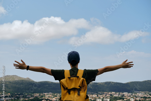 Travel and adventure concept. A young woman with yellow backpack stays in old town Bar in Montenegro. Girl with short hair and cap raised arms up in different directions, as if embracing whole world