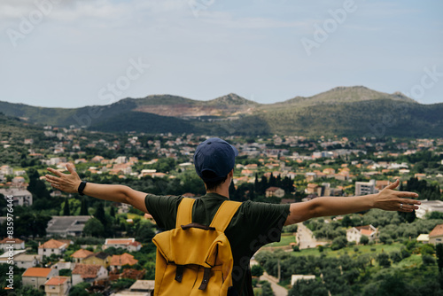 Travel and adventure concept. A young woman with yellow backpack stays in old town Bar in Montenegro. Girl with short hair and cap raised arms up in different directions, as if embracing whole world