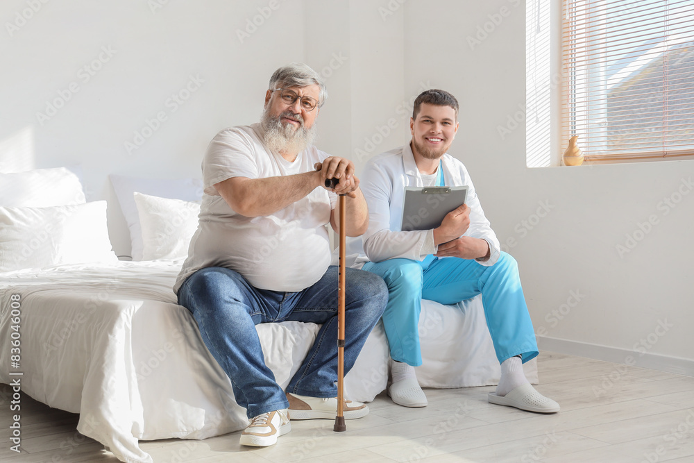 Obraz premium Young caregiver and senior man with stick sitting in bedroom