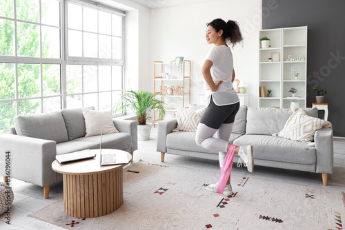 Sporty African-American woman with laptop and resistance band exercising at home