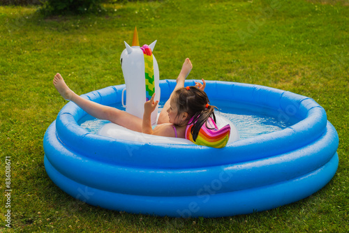 A girl child bathes with a rubber unicorn in an inflatable pool in nature on a hot summer day.