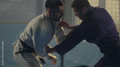 Slow-motion shot of barefoot martial arts athlete in blue kimono lifting his opponent up and throwing him down on mat during jujitsu practice in gym