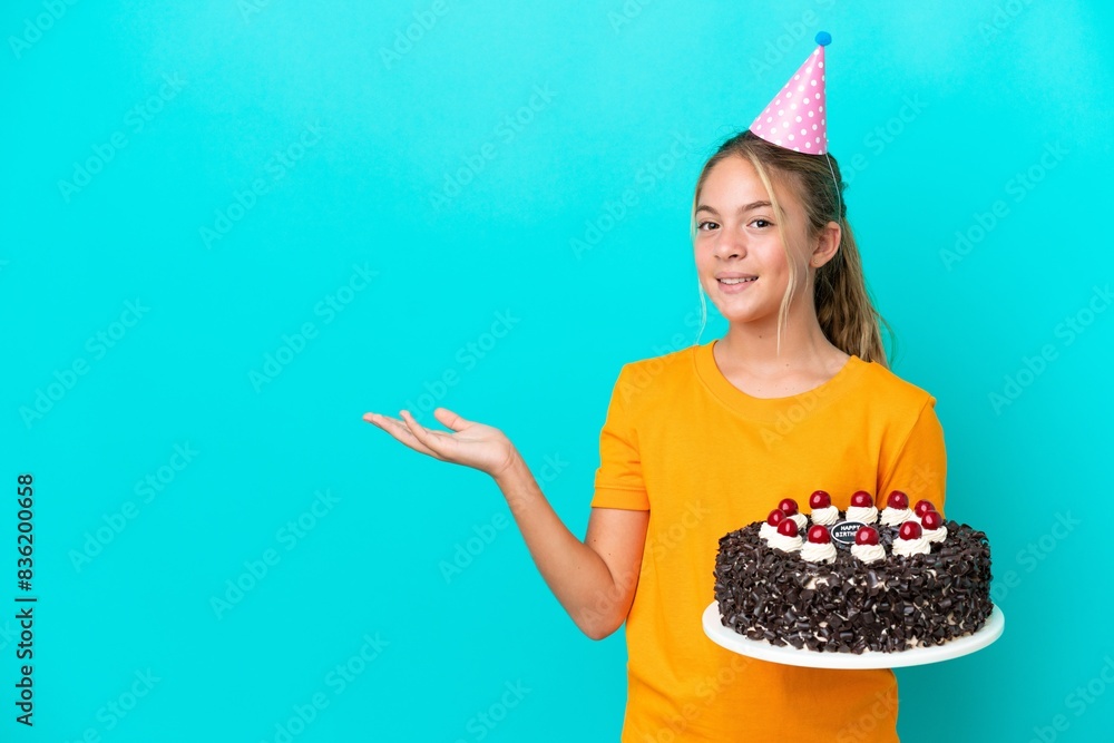 Little caucasian girl holding birthday cake isolated on blue background extending hands to the side for inviting to come Little caucasian girl holding birthday cake isolated on blue background extending hands to the side for inviting to come