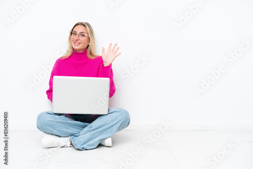 Young caucasian woman with laptop sitting on the floor saluting with hand with happy expression
