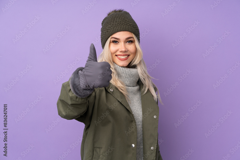 Teenager blonde girl with winter hat over isolated purple background with thumbs up because something good has happened Teenager blonde girl with winter hat over isolated purple background with thumbs up because something good has happened