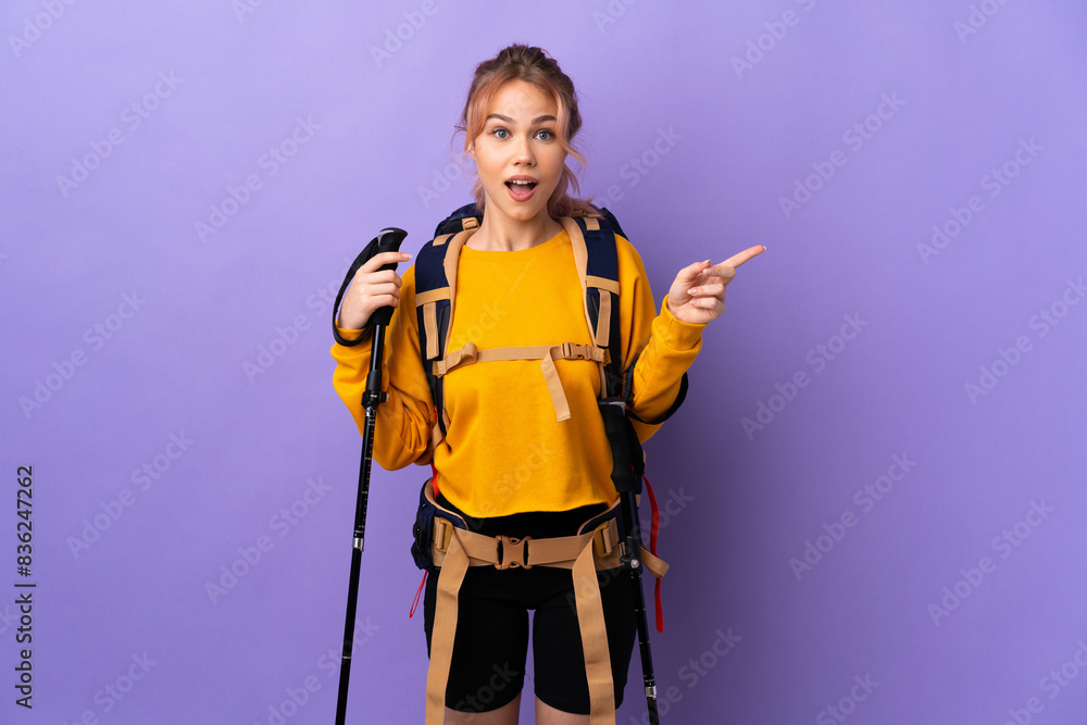 Teenager girl with backpack and trekking poles over isolated purple background surprised and pointing finger to the side Teenager girl with backpack and trekking poles over isolated purple background surprised and pointing finger to the side