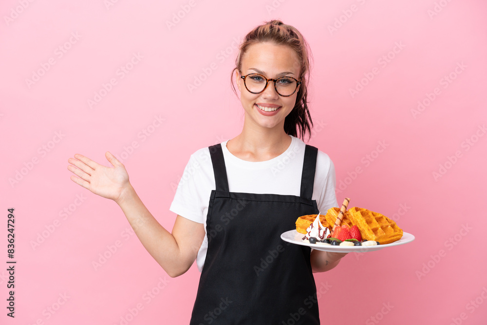 Restaurant waiter Russian girl holding waffles isolated on pink background extending hands to the side for inviting to come Restaurant waiter Russian girl holding waffles isolated on pink background extending hands to the side for inviting to come