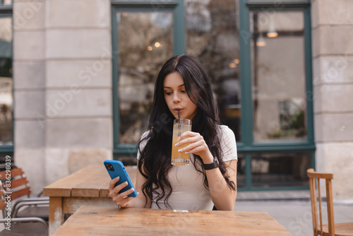 Young beautiful woman holding cocktail lemonade glass and looking at smartphone while sitting at cafeteria. Happy university student girl using mobile phone. Teen girl drink fresh cocktail and smiling