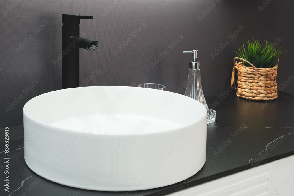 Fototapeta premium A white sink with a black faucet sits on a black countertop. A potted plant sits next to the sink, and a glass is on the counter