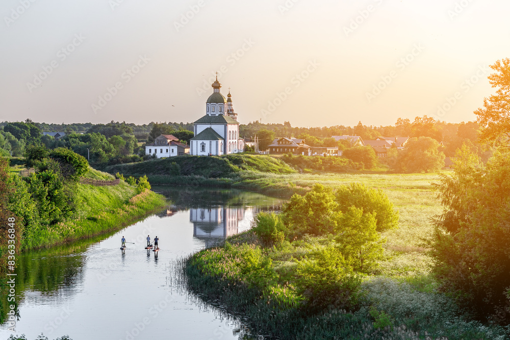 Summer landscape in Suzdal. Church of Elijah the Prophet on the bank of the river Kamenka. Tourists on SUP boards float on the river. Russia, Vladimir region, Golden Ring of Russia Summer landscape in Suzdal. Church of Elijah the Prophet on the bank of the river Kamenka. Tourists on SUP boards float on the river. Russia, Vladimir region, Golden Ring of Russia