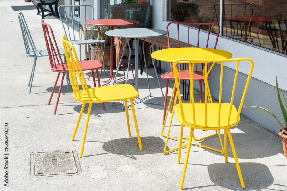 Multiple empty bright grey, yellow, and red colored metal round tables and highback chairs at a bistro cafe on a restaurant's sidewalk patio. The restaurant has large glass windows and a white wall. Multiple empty bright grey, yellow, and red colored metal round tables and highback chairs at a bistro cafe on a restaurant's sidewalk patio. The restaurant has large glass windows and a white wall.