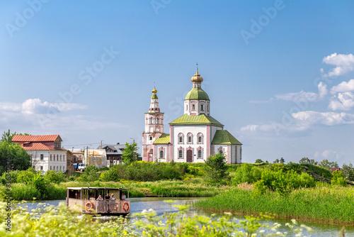 SUZDAL, RUSSIA - A beautiful landscape of Suzdal with a view from the high bank of the Kamenka River on Pushkarskaya Street to the river and a pleasure boat with people photo