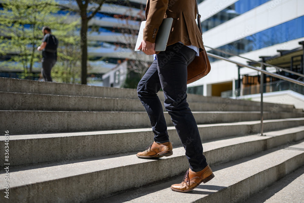 Low section of male commuter with shoulder bag and laptop walking up on staircase in modern city Low section of male commuter with shoulder bag and laptop walking up on staircase in modern city