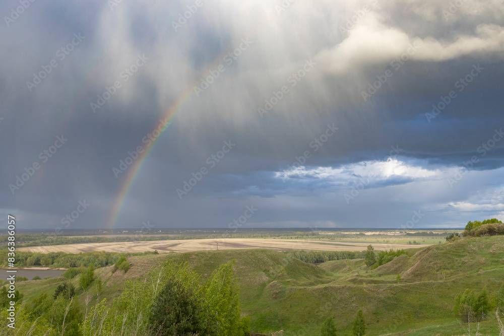 A tranquil rural landscape under a dramatic sky, where a rainbow stretches across the horizon, casting a spectrum of colors over the rolling hills and agricultural fields. A tranquil rural landscape under a dramatic sky, where a rainbow stretches across the horizon, casting a spectrum of colors over the rolling hills and agricultural fields.