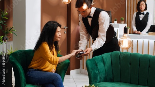Asian client paying for coffee served in lounge area, using pos terminal payment to buy drink from hotel bar. Traveler making transaction with credit card upon her arrival at holiday retreat.