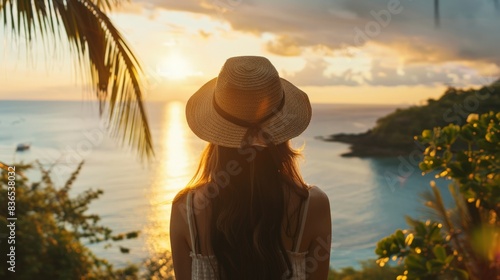 A young woman  adorned with a hat  overlooks a calm sea in a lush tropical setting. The wide shot captures the serene vibe  bathed in the warm glow of golden hour light.