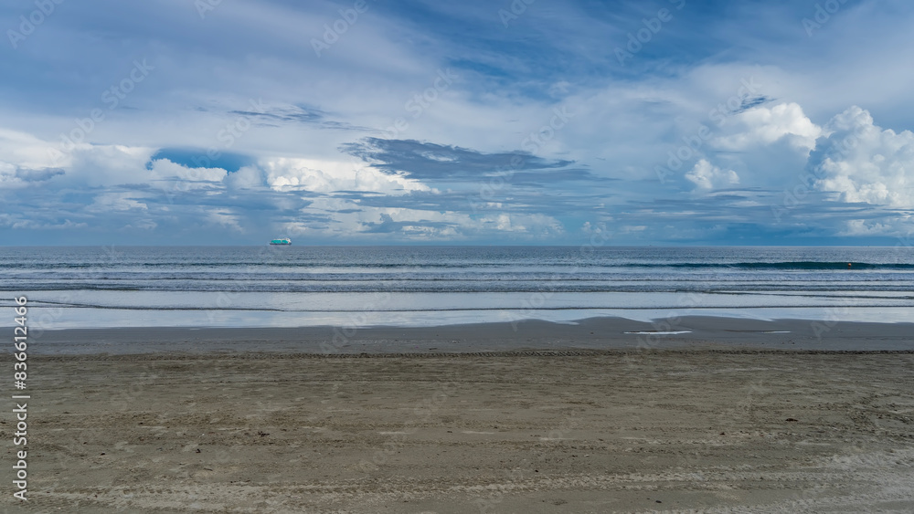 Fototapeta premium Morning on the ocean coast. Turquoise waves calmly roll towards the shore, spreading along the beach. There are tracks of cleaning machines in the sand. A ship is on the horizon. Clouds, blue sky