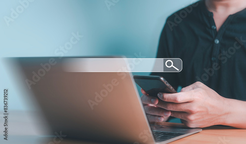 A person holding a smartphone while using a laptop, symbolizing online search and multitasking in a digital workspace.