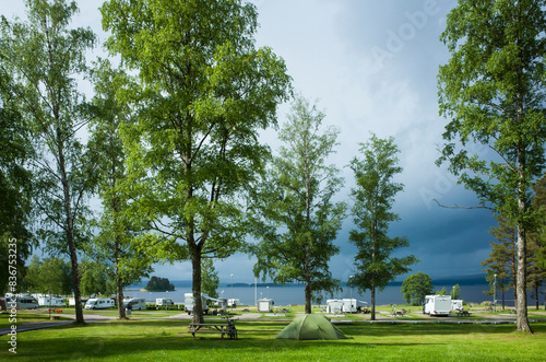 Campsite with green tent on grass, Campervans parked behind tall trees by the lake, Gray rainy sky, Sun shining, Travel in Värmland, Glafsfjorden lake, Sweden