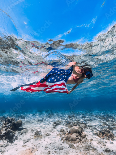 Woman swims with US flag underwater in tropical blue ocean. Independence day concept.