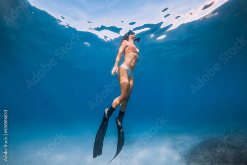 Freediver girl with fins swims underwater over sandy sea in blue ocean.