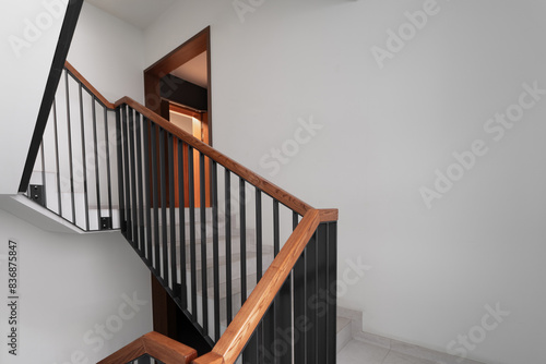 A contemporary staircase design featuring a sleek wooden handrail and black metal balusters, leading up to a well-lit hallway with minimalistic decor and clean lines.