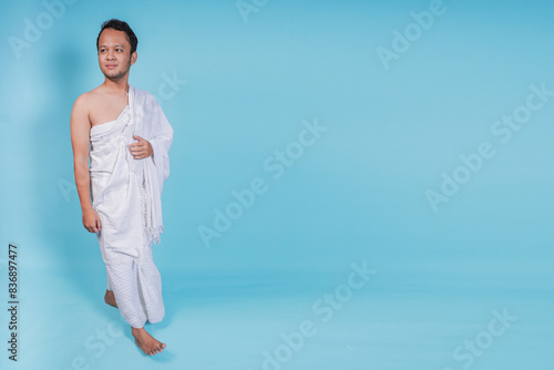 Handsome young Asian Muslim man wearing ihram walking over isolated by blue background. Hajj concept. © Reezky